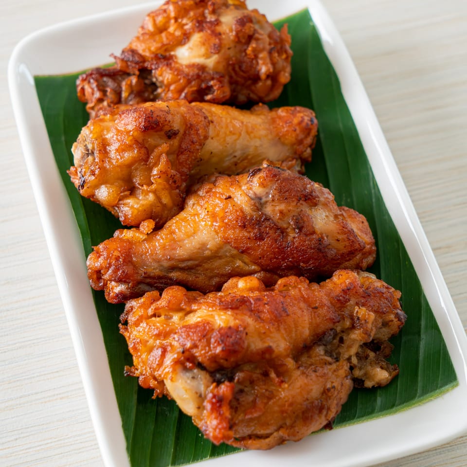 Four crispy, golden-brown fried chicken pieces on a white rectangular plate with green banana leaves.